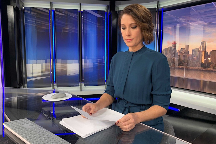 Tamara Oudyn sits at the news desk in the television studio looking down at white paper.