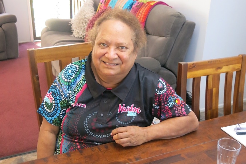 An Indigenous woman sitting at a wooden dining table, with a brown couch in the background.