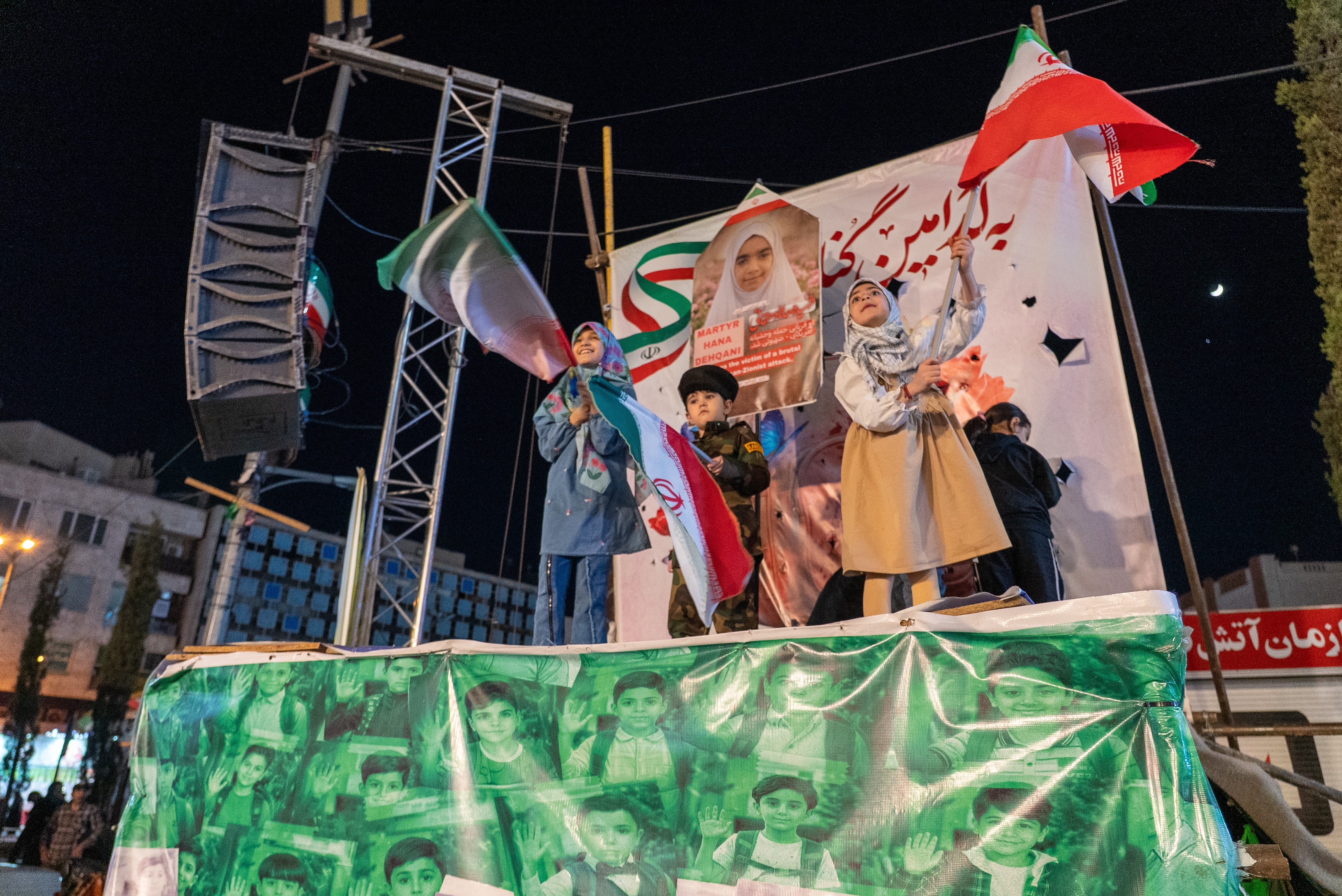 Two girls and a young boy standing on a stage waving Iranian flags.