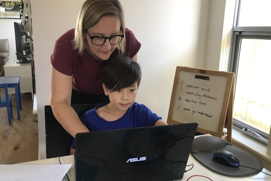 A mother leaning over her son to help him work on a laptop