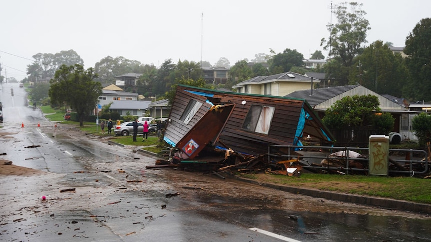 Cloudy image of house frame warped and moved by floods, debris on road and around base of structure. 