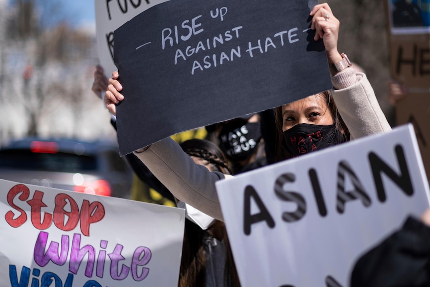 An Asian woman in face mask surrounded by signs lifts a 'Rise Up Against Hate' sign at outdoor rally
