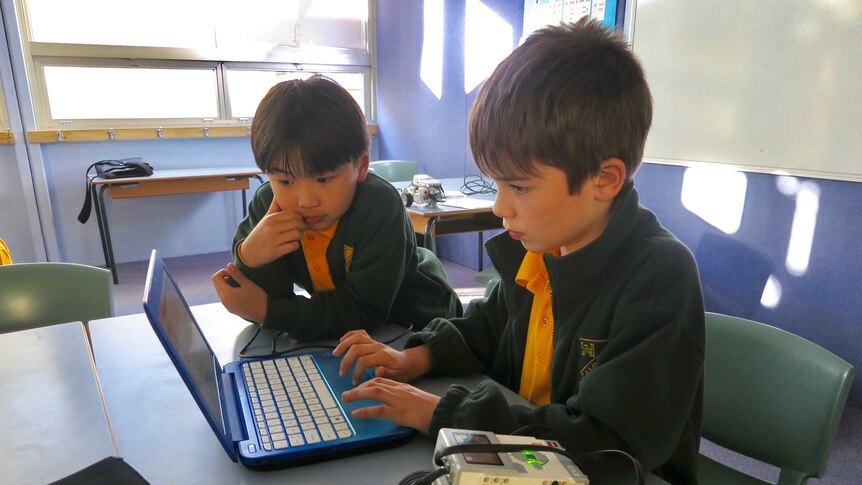 Two male primary school students look at a compute screen in a classroom with a small robot on the table near them