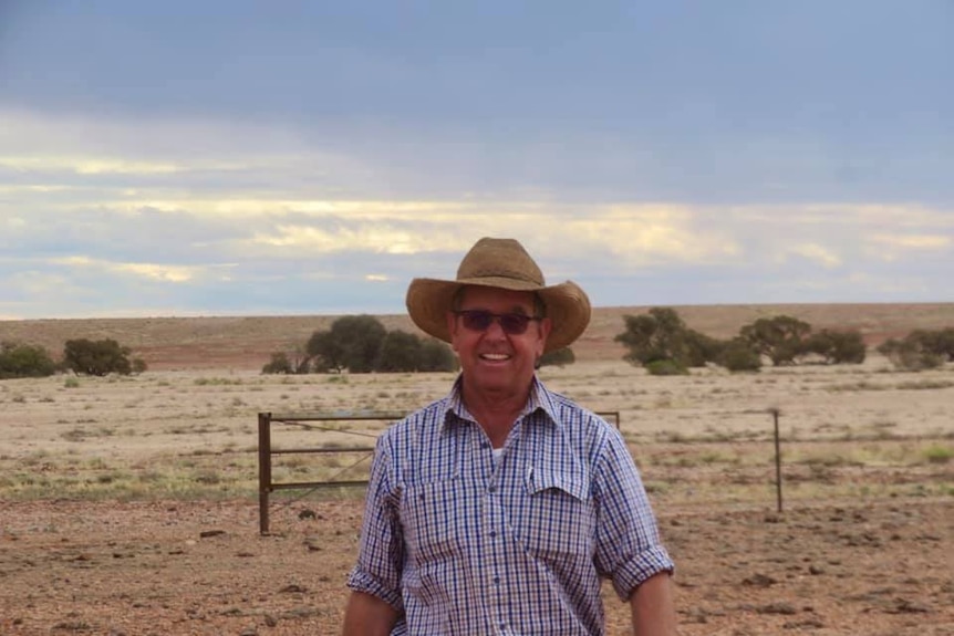 A grinning man in a hat and sunglasses stands in a desert paddock with fencing behind behind him.