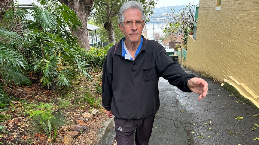 A man with grey hair and black tracksuit swings his arm out in front walking on footpath 