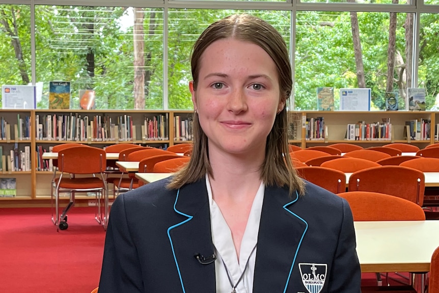 Freya sits in a classroom. She wears a blue blazer and white shirt. There is a bookshelf behind her.