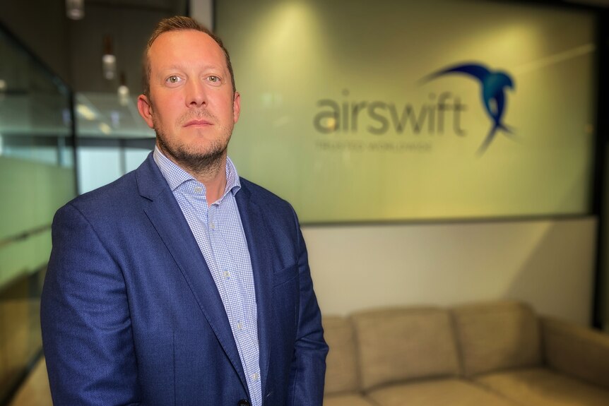 A serious middle-aged man in blue suit, white and blue patterned shirt in front of a wall with airsiwft and its logo, a couch.
