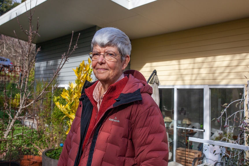 An elderly woman stands outside her home in the garden