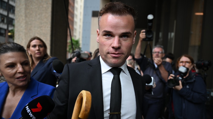 A man wearing a suit and tie carrying an umbrella surrounded by journalists