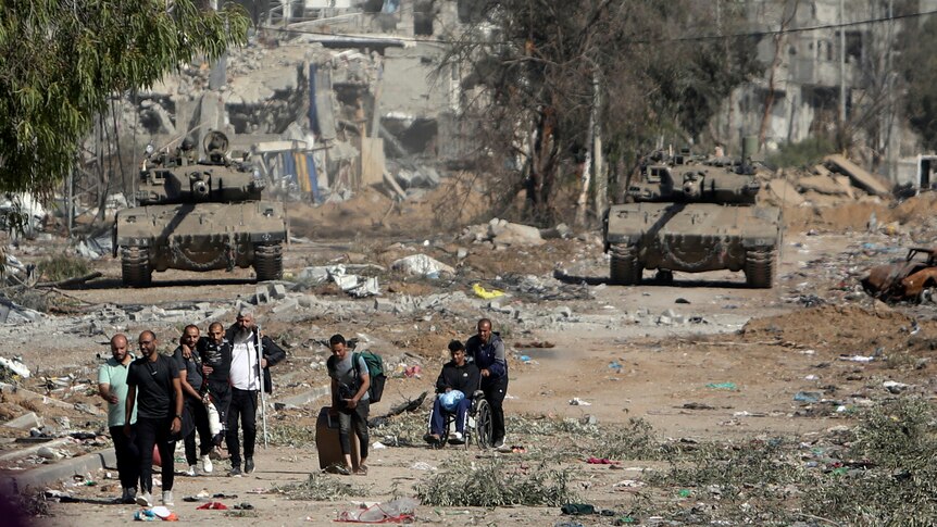 Two tanks sit amid damaged building facing a group of people leaving down a dirt road
