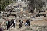 Two tanks sit amid damaged building facing a group of people leaving down a dirt road