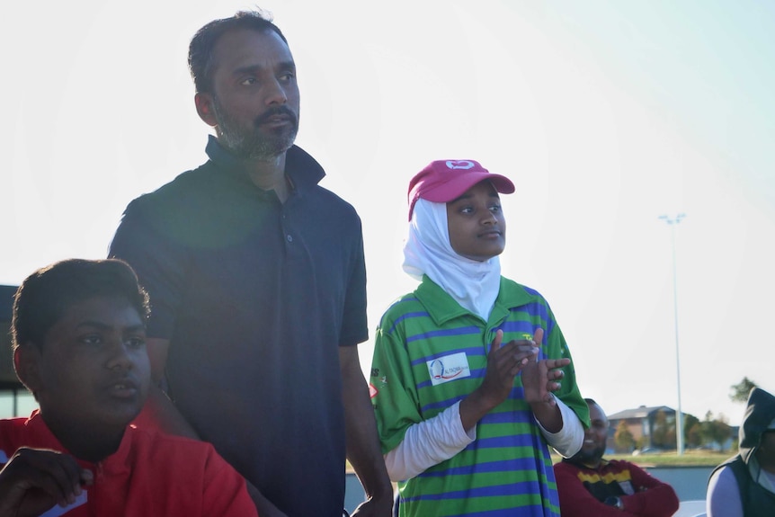 A man with short hair and a beard stands next to his daughter, in a green and blue cricket uniform.