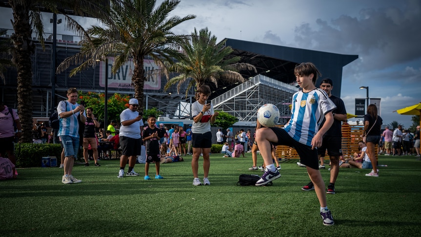 A child wearing a blue and white striped soccer jersey balances a ball on his knee while adults watch on