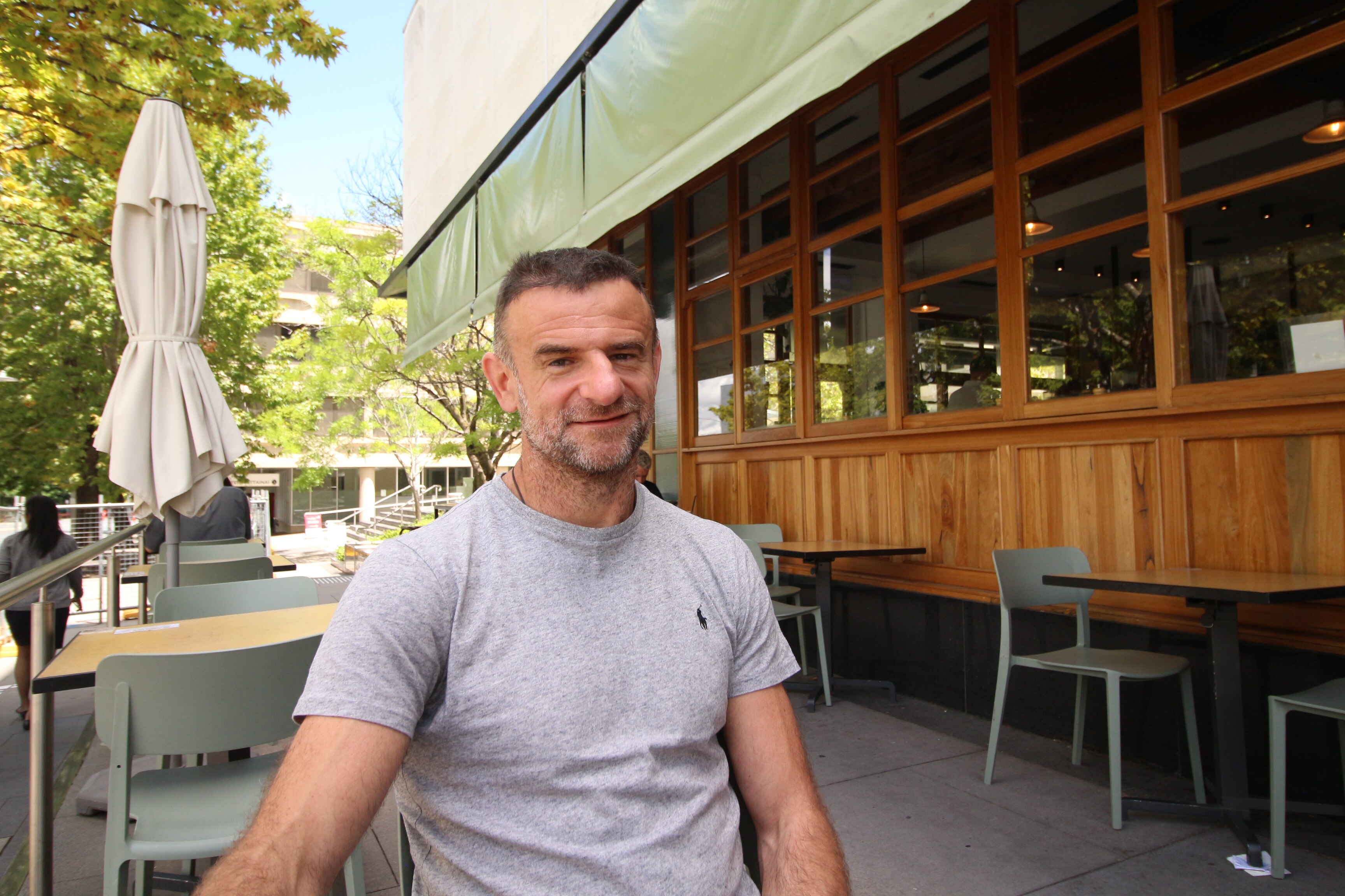 Un hombre con cabello gris se sienta afuera de un café vacío que se ve serio.