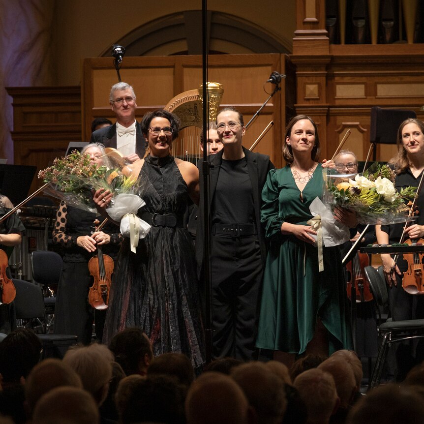 Claire Edwardes, Elena Schwarz and Anne Cawrse stand on stage in front of the Adelaide Symphony Orchestra.