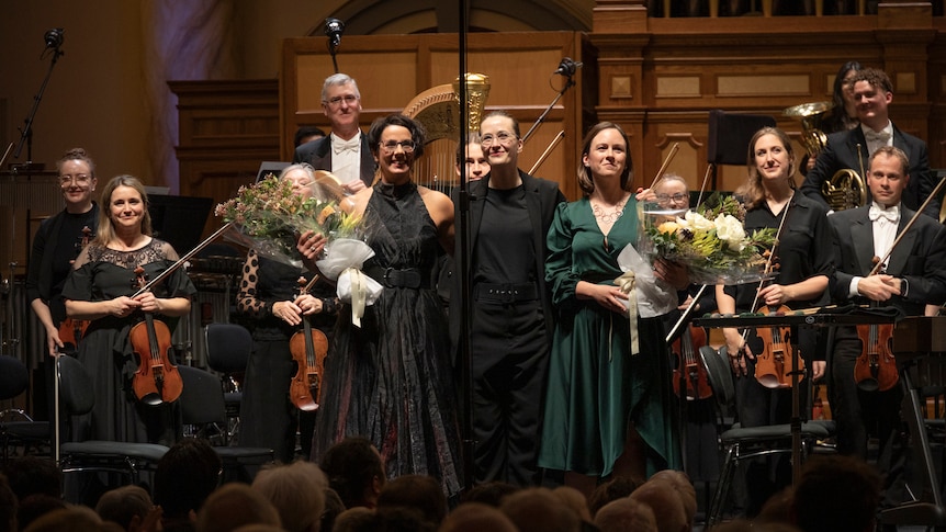 Claire Edwardes, Elena Schwarz and Anne Cawrse stand on stage in front of the Adelaide Symphony Orchestra.