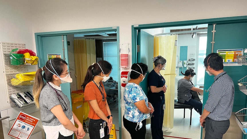 Hospital staff stand outside of a room as Dr Michael Lam plays cello to a patient