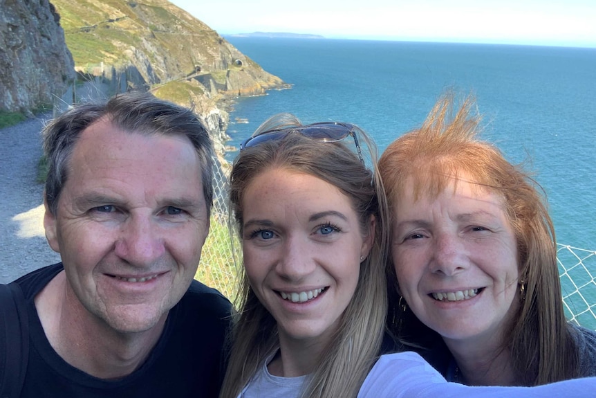 A young woman with her father to the left and mother to the right. Behind them is a cliff and the ocean.