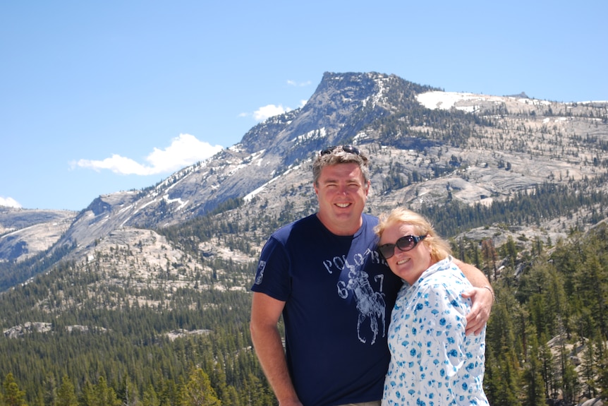 A middle-aged man and woman stand in front of a mountain with snow.