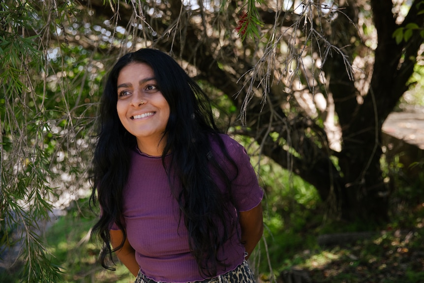 A woman smiling, standing in front of a tree.