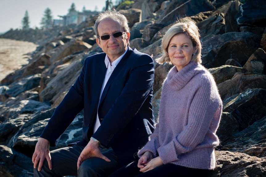 A man in a suit and glasses sits next to a woman wearing a purple sweater, both sit on rocks at the beach