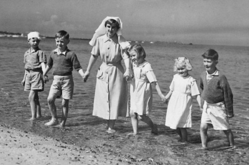 Black and white archival photo of a nurse holding hands with 5 children at a beach