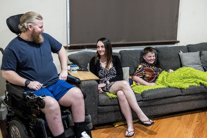 A paraplegic man in a wheelchair next to a woman and child sitting relaxed on a couch.  