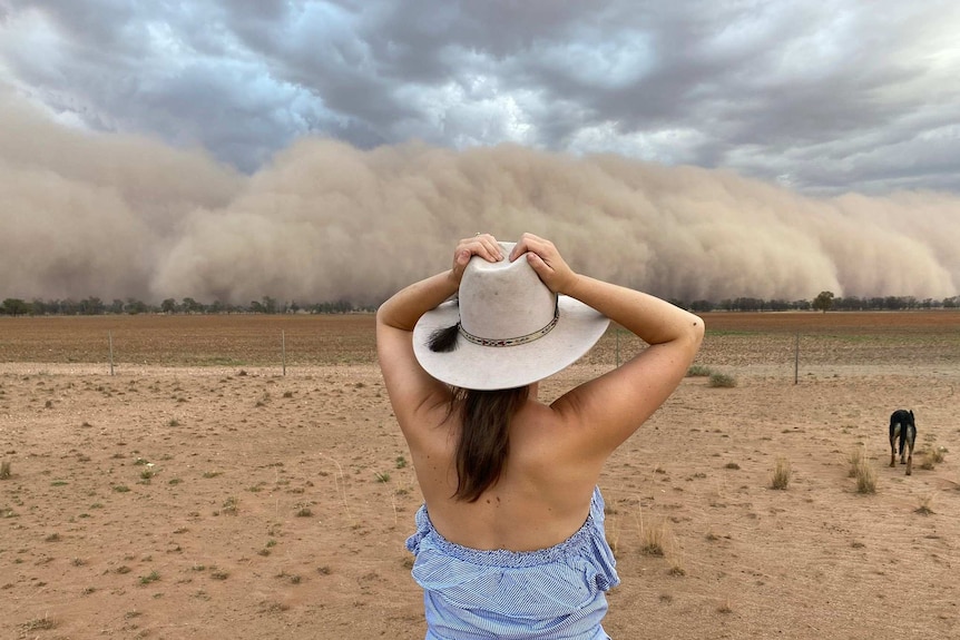 A woman in an Akubra hat with a dog standing in a very dry paddock as a wall of dust approaches.