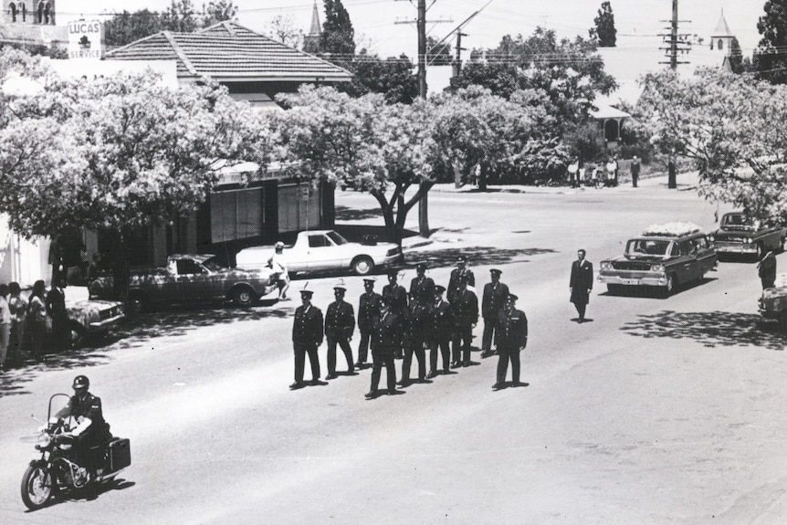 Black and white image of a police funeral, officers march the street followed by Tracker Riley in a  hearse