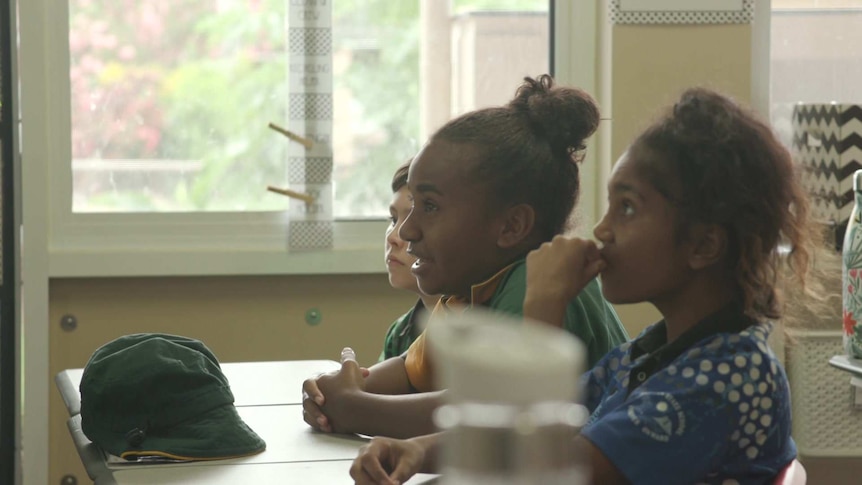 Students sit at desk in class, listening to a teacher off-camera