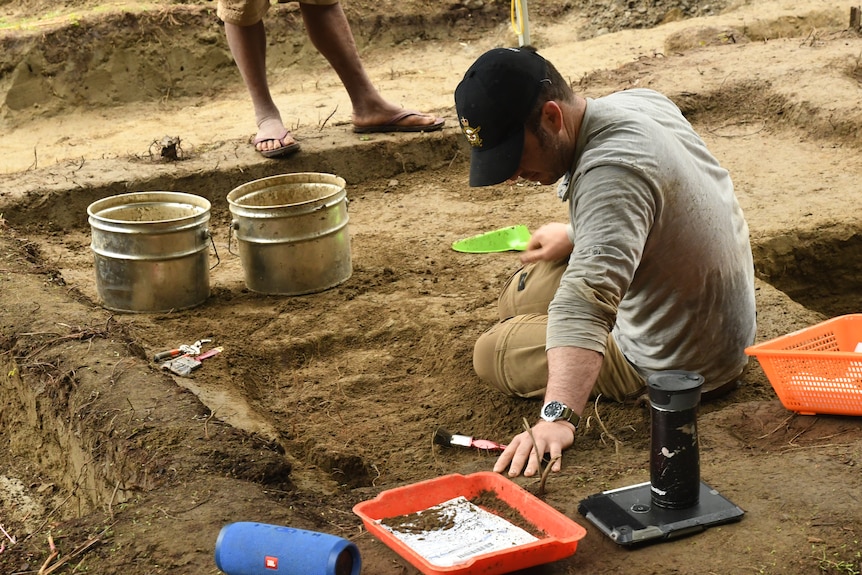 A man wearing a long-sleeved grey shirt sits in the dirt with a brush in one hand and dust pan in the other, buckets nearby