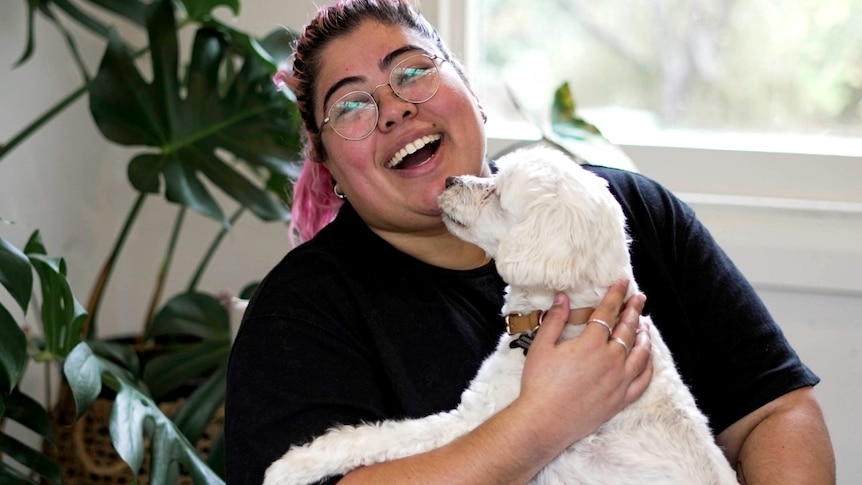 A young woman with brown and pink hair cuddles a small white dog