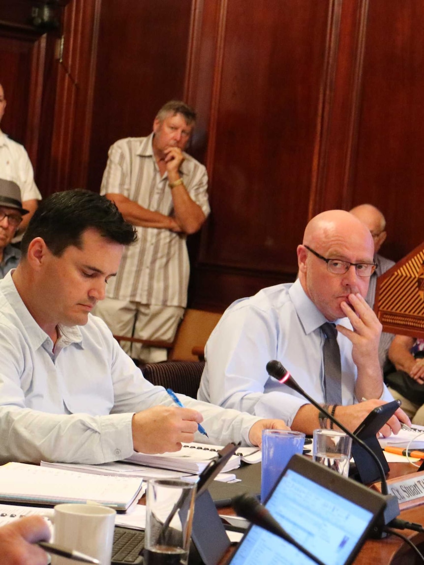 Four men at a table with a small with microphones with a crowd behind them looking on