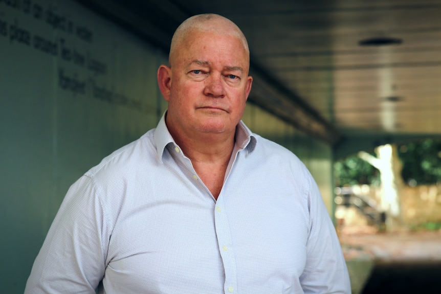 A large, older man stands in front of a building, wearing a light blue shirt.