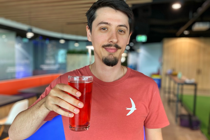 Adam Chalmers holds up a glass of red liquid - possibly kombucha - while standing in an office.
