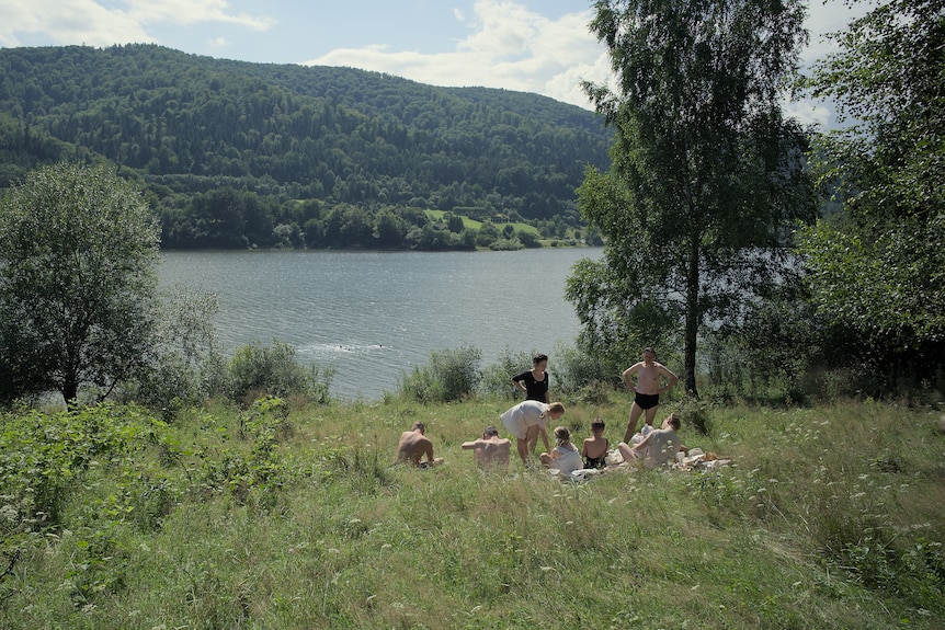 A film still of a group of people in 40s-style dress sitting in the grass by a river.