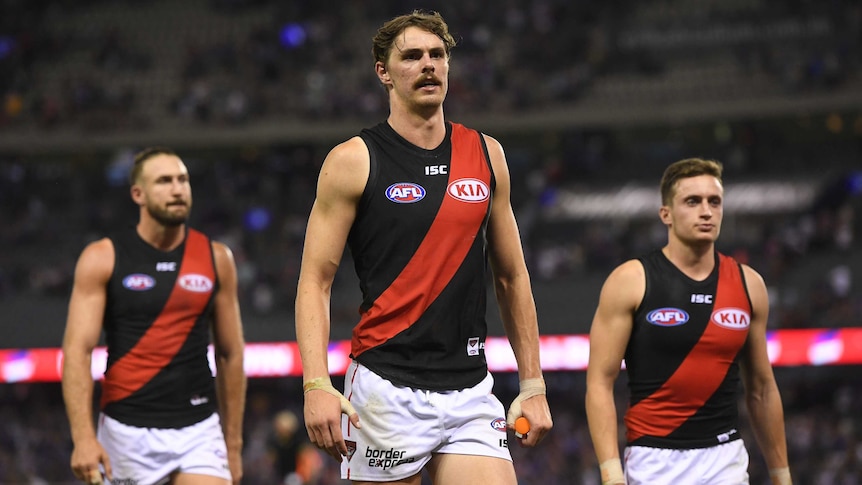 An Essendon AFL player walks off the field with two teammates walking behind him.