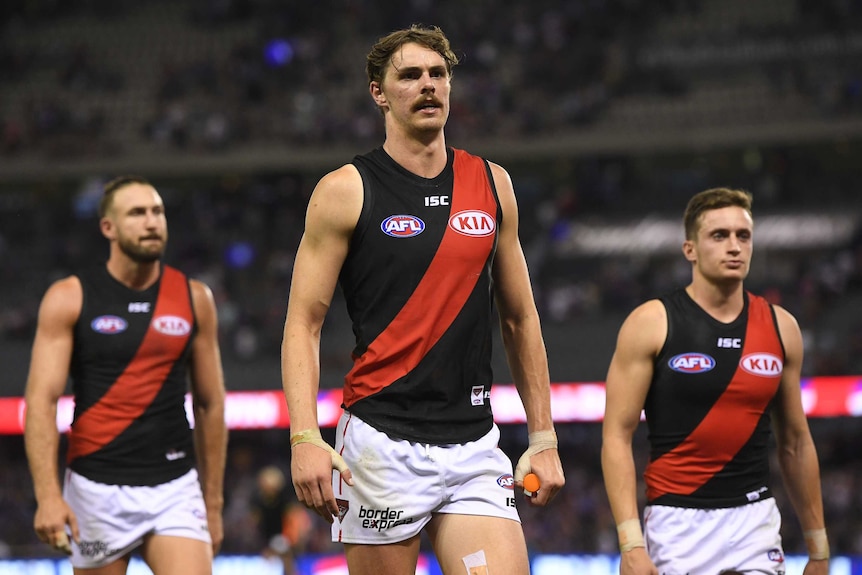 An Essendon AFL player walks off the field with two teammates walking behind him.