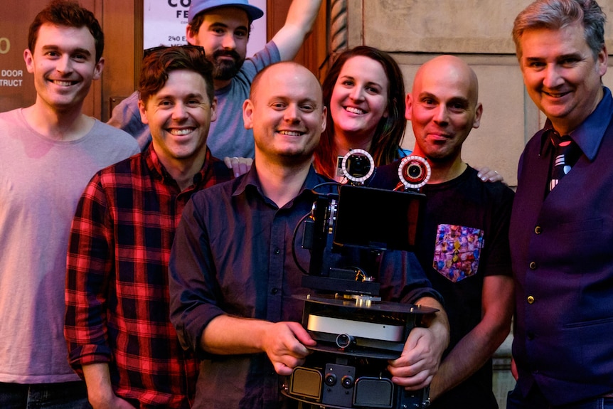 Six men and one woman stand smiling behind a robot named LOL-BOT