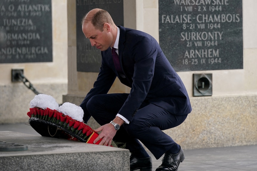 Prince William crouches as he places a wreath of read an white flowers at the base of an ornate stone monument