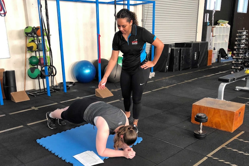 A woman in black gym gear stands over her client who is in a plank position.
