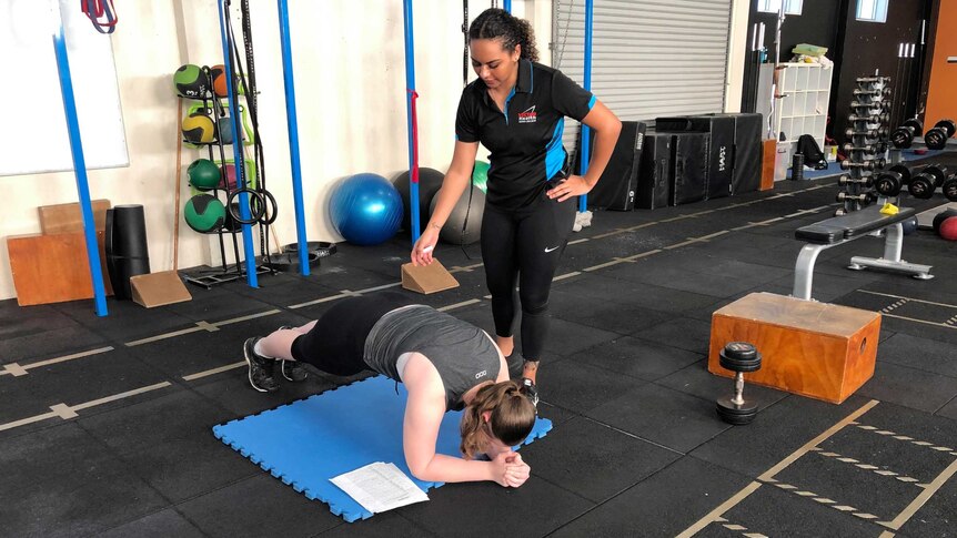 A woman in black gym gear stands over her client who is in a plank position.