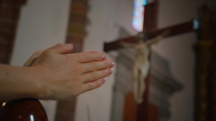 close up of hands in prayer in the inside of a church, statue depicting Jesus hanging on the cross in the background