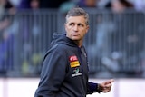 A medium close up shot of Fremantle coach Justin Longmuir wearing a grey hoodie walking on the field and looking to the right.