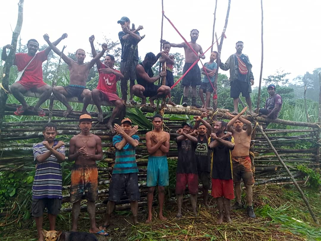 A group of men cross or raise their arms while standing in front of a makeshift blockade made of timber.
