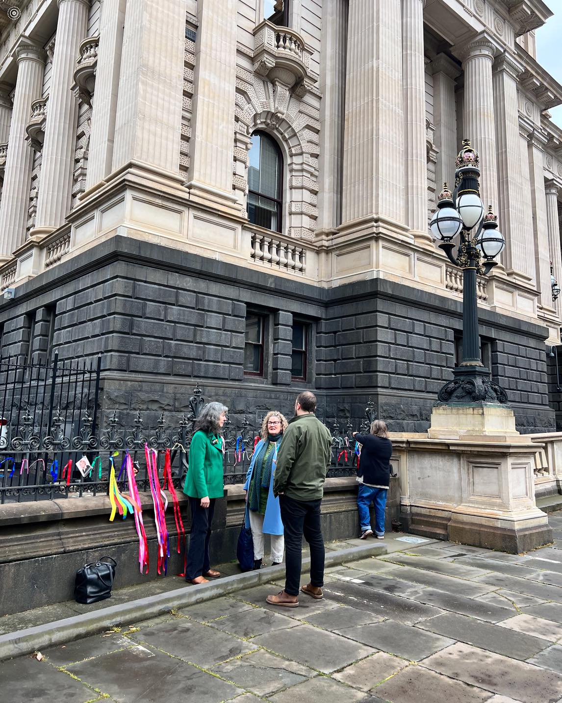 Three people stand outside Victoria's Parliament House, an ornate cream and black building.
