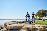 Two people on e-scooters pose by a rocky outcrop near a beach