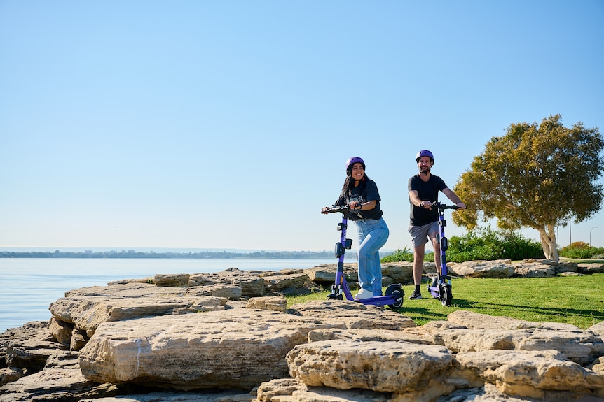 Two people on e-scooters pose by a rocky outcrop near a beach