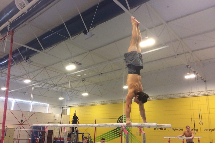 A member of the Australian men's gymnastics team suspends himself upside down on parallel bars as his team mates watch on.