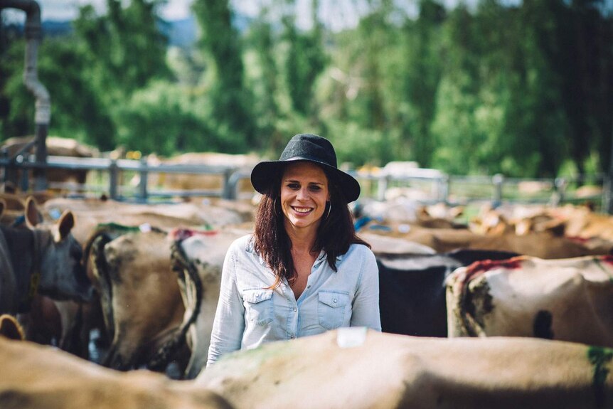 A woman with a broad-brim hat smiles as the camera surrounded by jersey cattle